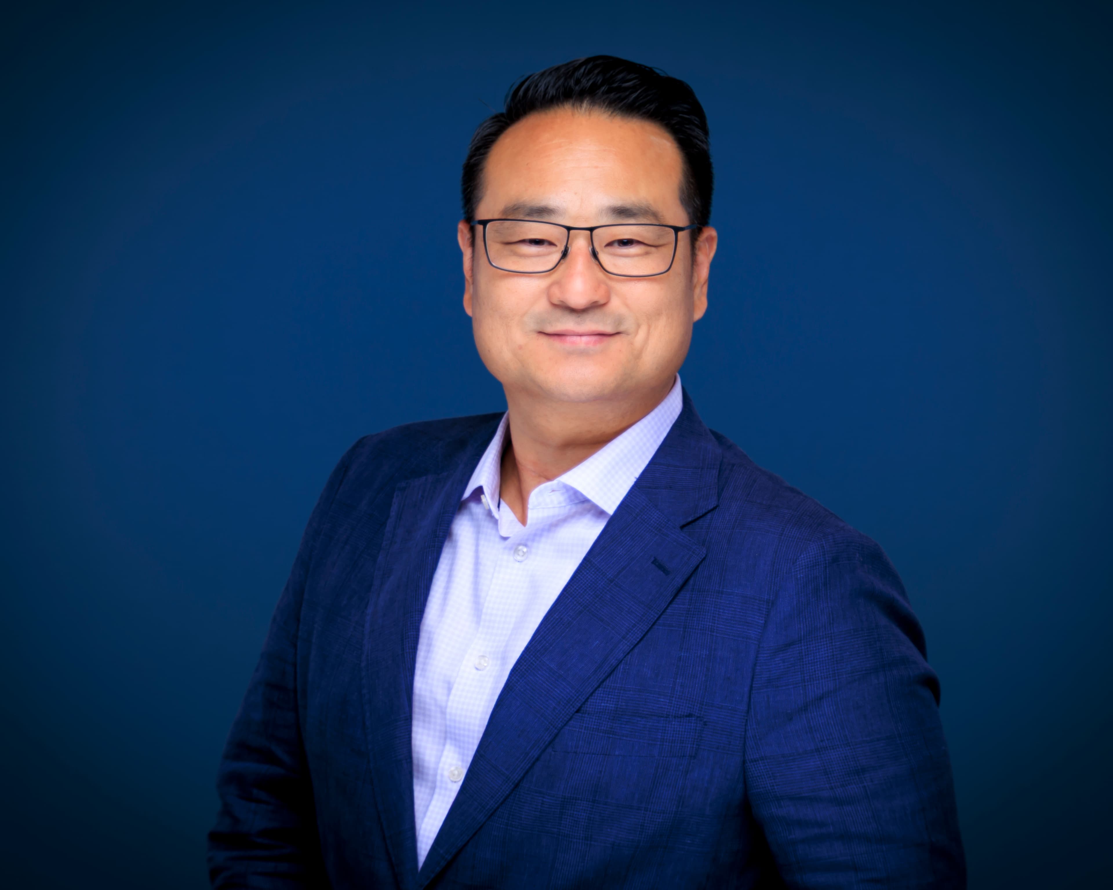Smiling Asian man with glasses wearing a blue blazer against a dark blue background.