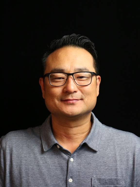Smiling Asian man wearing glasses and a grey collared shirt against a black background.